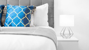 En pænt redt seng med en pude i hvidt og blåt mønster, en ensfarvet hvid pude og en geometrisk lampe på et hvidt natbord.