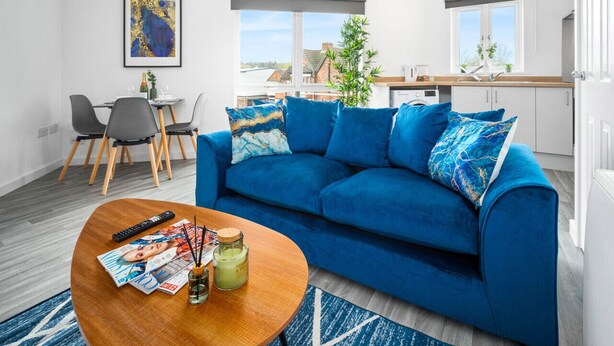 Uma sala de estar moderna com um sofá azul, uma mesa de centro de madeira com revistas e vista para a cidade através de grandes janelas.