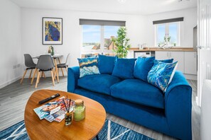 Una sala de estar moderna con un sofá azul, una mesa de centro de madera con revistas, y vistas a la ciudad a través de amplias ventanas.