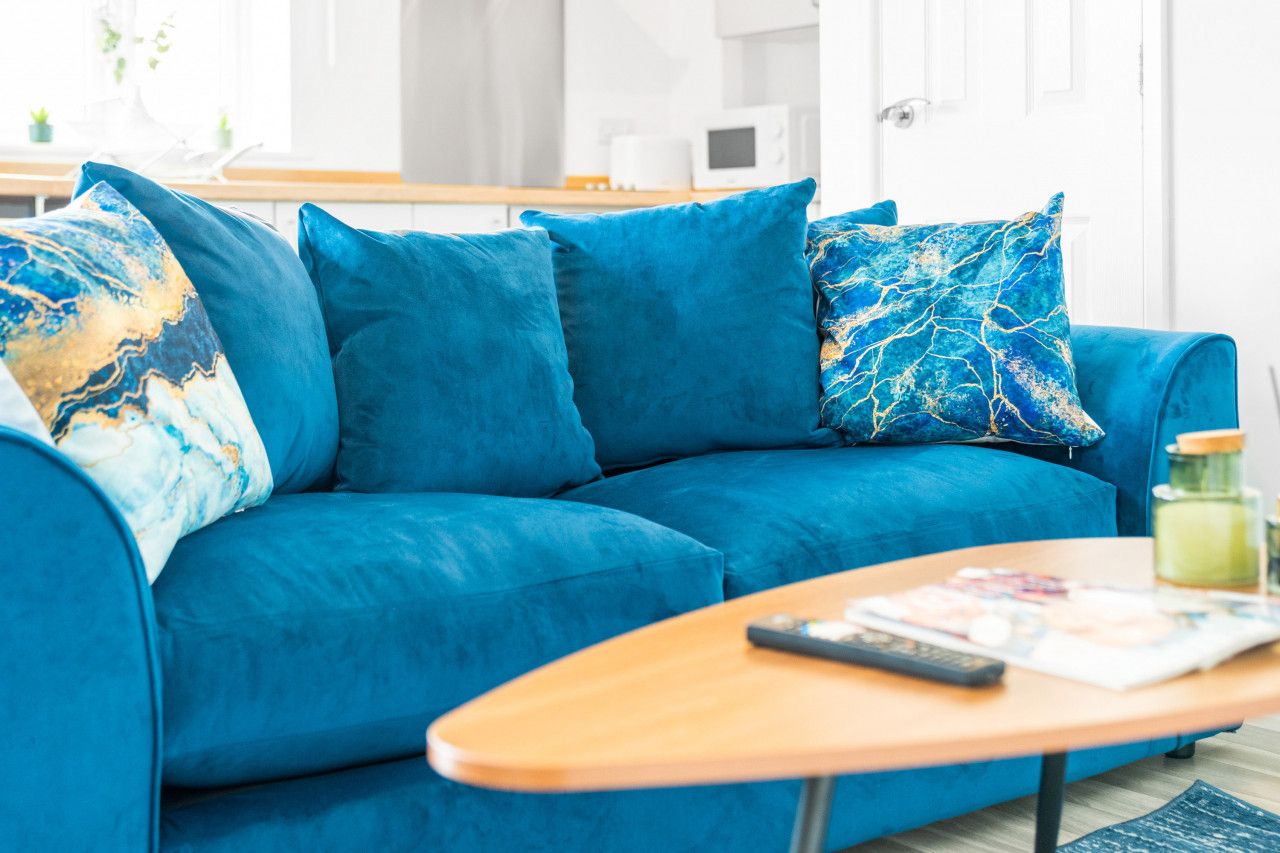 Un divano blu con cuscini coordinati, un tavolino in legno con un telecomando e una rivista, e una cucina sullo sfondo.