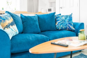 Ein blaues Sofa mit passenden Kissen, ein Holztisch mit Fernbedienung und Zeitschrift, und eine Küche im Hintergrund.