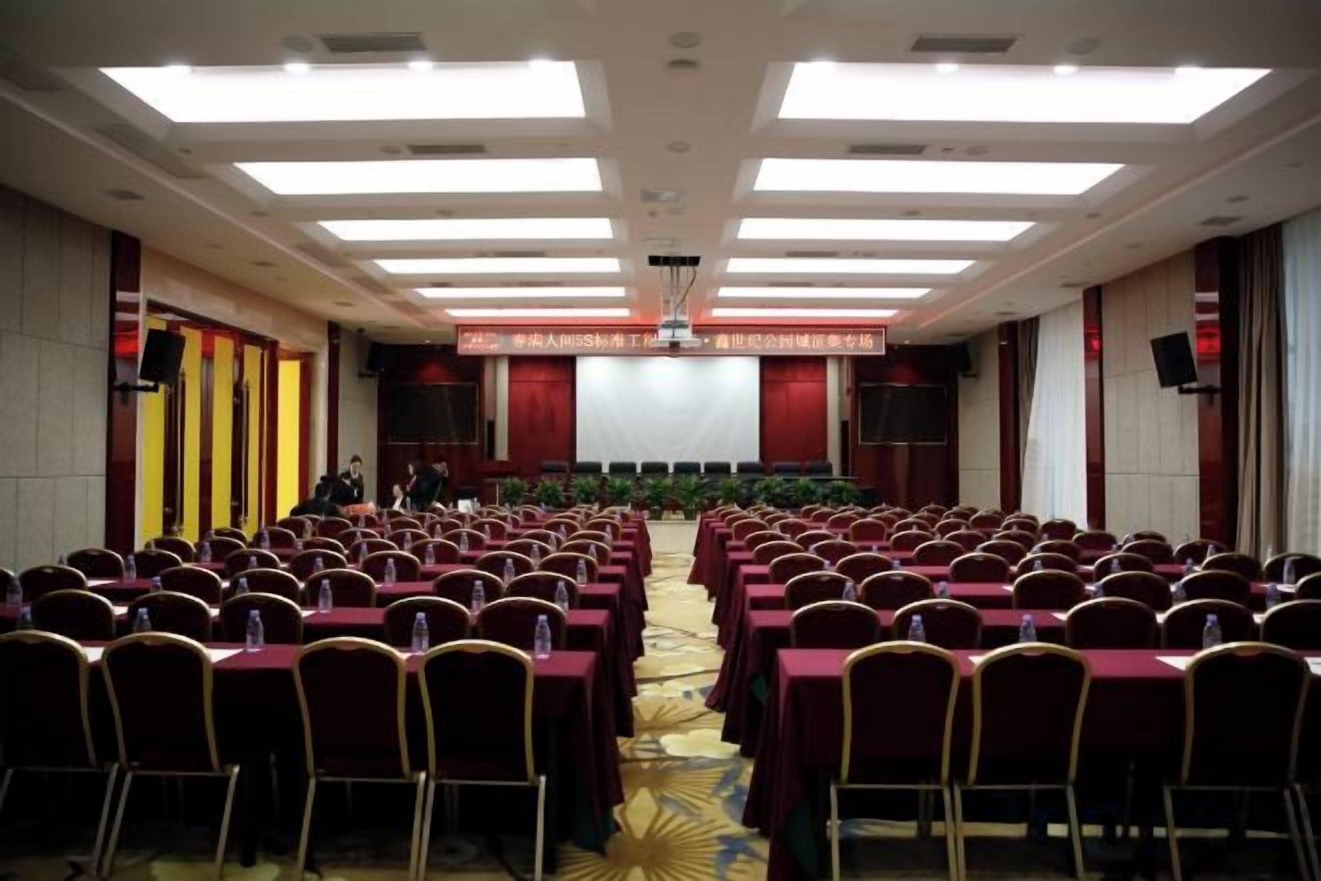 A conference room with rows of tables and chairs, a stage with a screen, and a carpeted floor.
