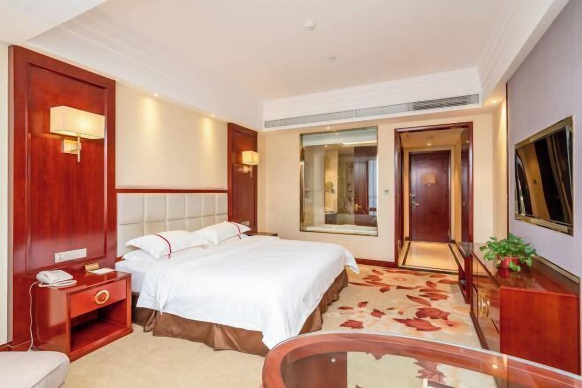 Habitación de hotel con una cama grande, una mesita de noche con lámpara, un espejo y vistas al pasillo.