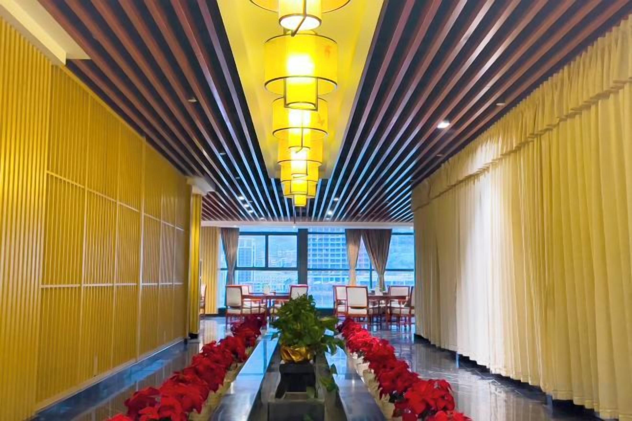 A hotel corridor with a central floral arrangement, a row of chairs, and a view of the cityscape through large windows.