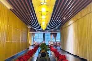 A hotel corridor with a central floral arrangement, a row of chairs, and a view of the cityscape through large windows.