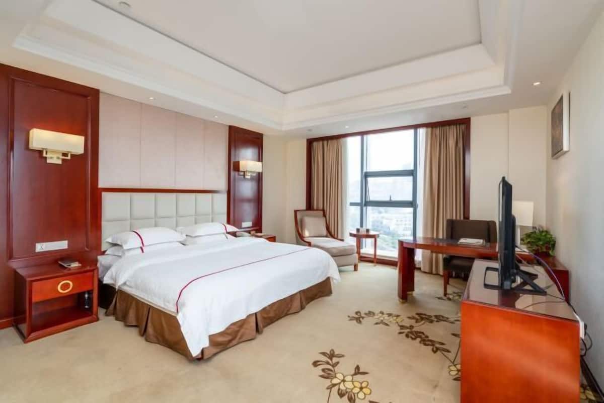 Habitación de hotel con una cama grande, un escritorio con televisor, una silla y un ventanal con cortinas.