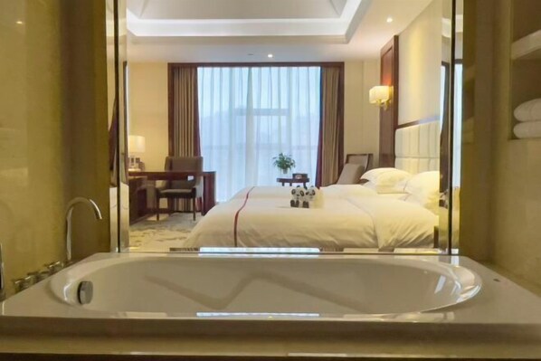 Ein Hotelzimmer mit einer großen Badewanne, einem Bett mit weißer Bettwäsche, einem Schreibtisch mit Stuhl und einem Fenster mit Vorhängen.