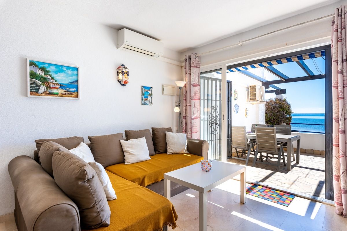 A modern living room with a sofa, a small table, and a view of the sea through a glass door.