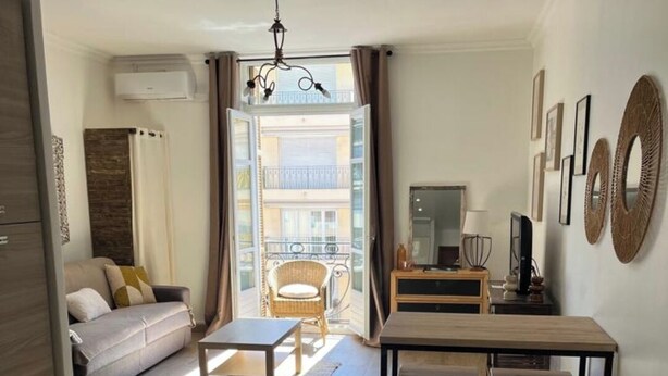 En moderne stue med sofa, spisebord med to stole, et lille bord og et fjernsyn. Der er en balkon med en stol og udsigt til bygninger.