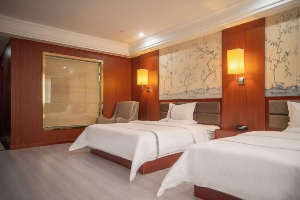 A hotel room with two beds, a large window with blinds, and wall-mounted lamps.