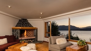 Una sala de estar acogedora con chimenea, muebles de madera y un amplio ventanal con vistas a un lago y montañas.