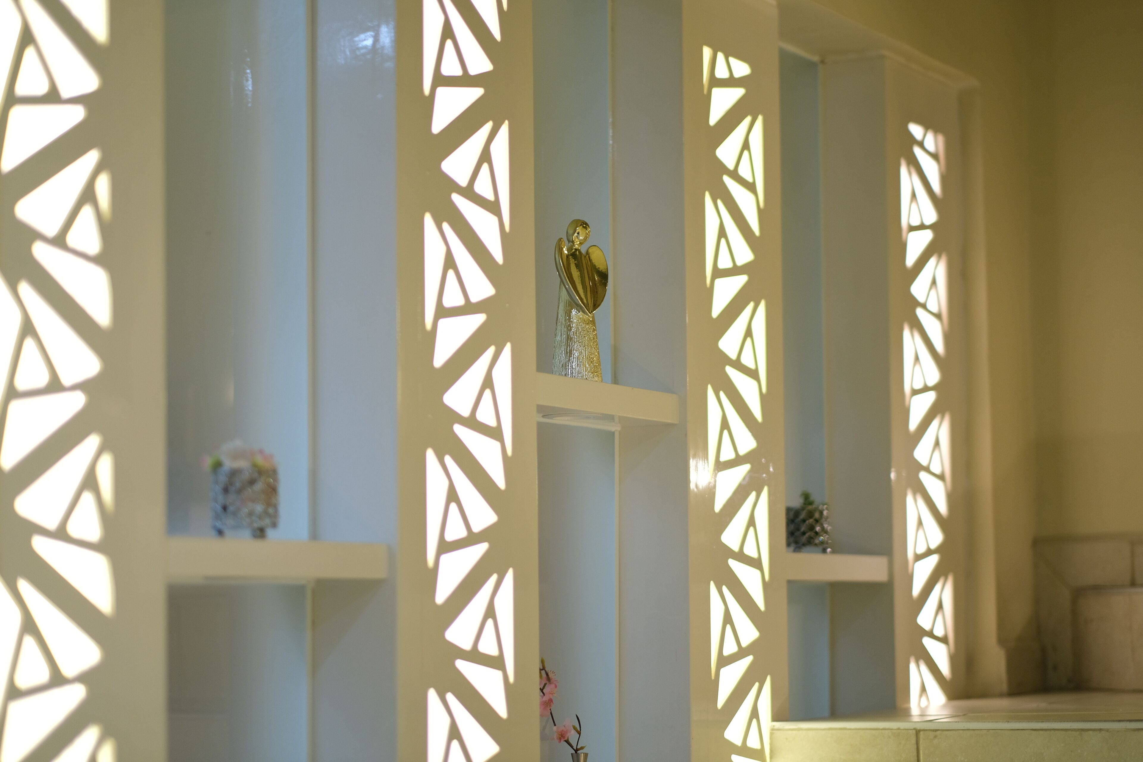 A room with a decorative partition featuring geometric cut-out patterns, a small decorative item on the lower shelf, and a vase with flowers.