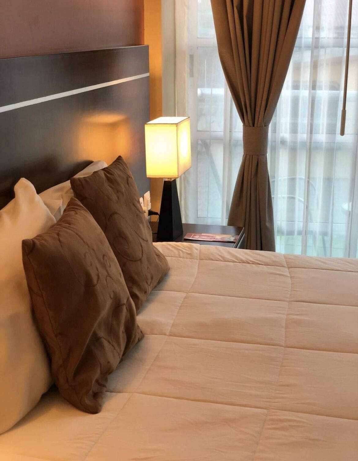 A hotel room with a neatly made bed, a bedside lamp, and a view of the city through sheer curtains.