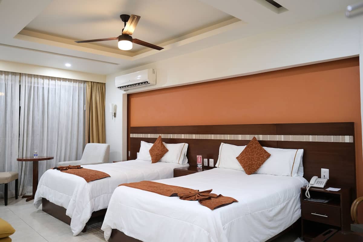A hotel room with two beds, a ceiling fan, an air conditioning unit, a bedside table with a phone, and a small table with a chair.