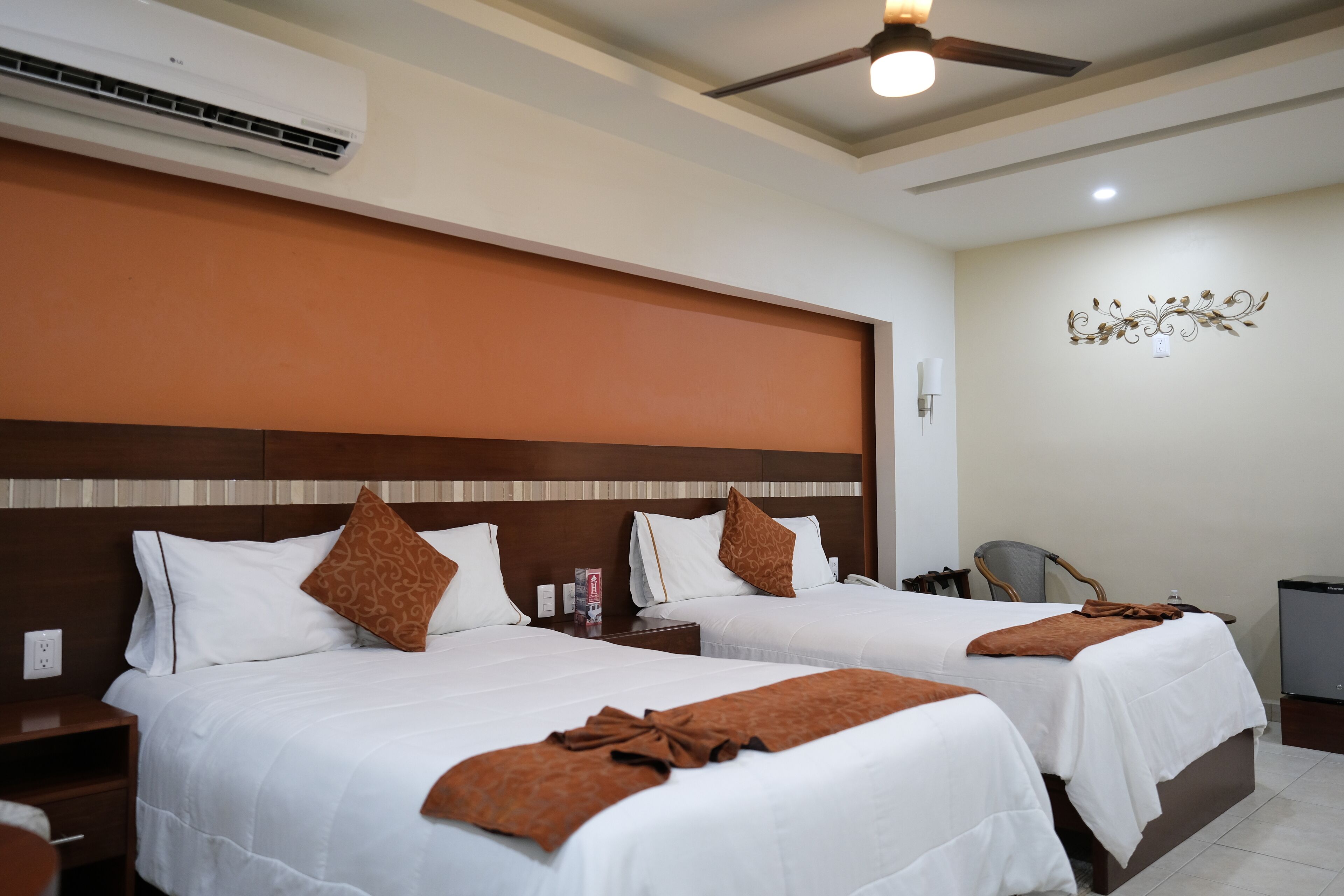 Habitación de hotel con dos camas, ventilador de techo, aire acondicionado, un adorno en la pared y televisión.
