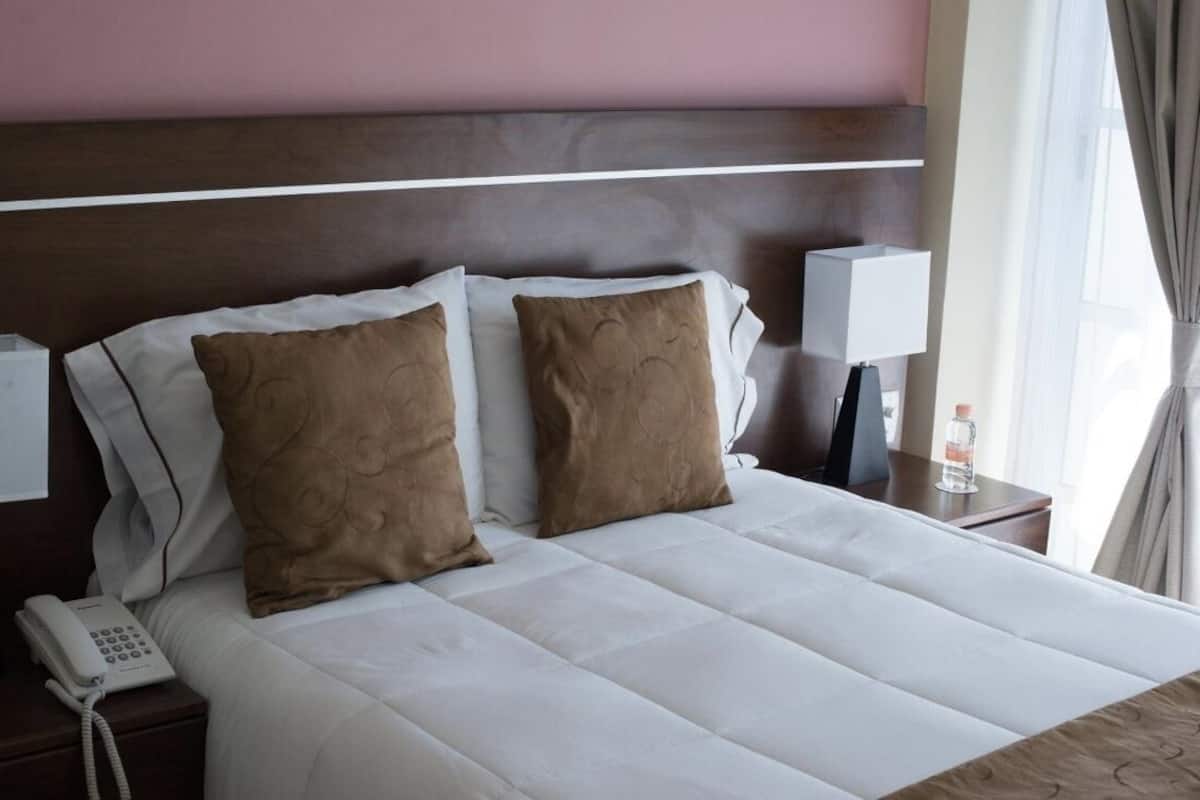 A hotel room with a large bed, two pillows, a bedside lamp, a phone, and a window with curtains.