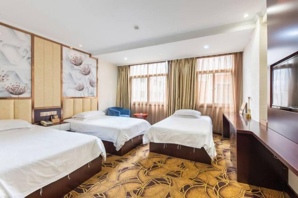 Habitación de hotel con tres camas, televisión, escritorio y un ventanal grande con cortinas.