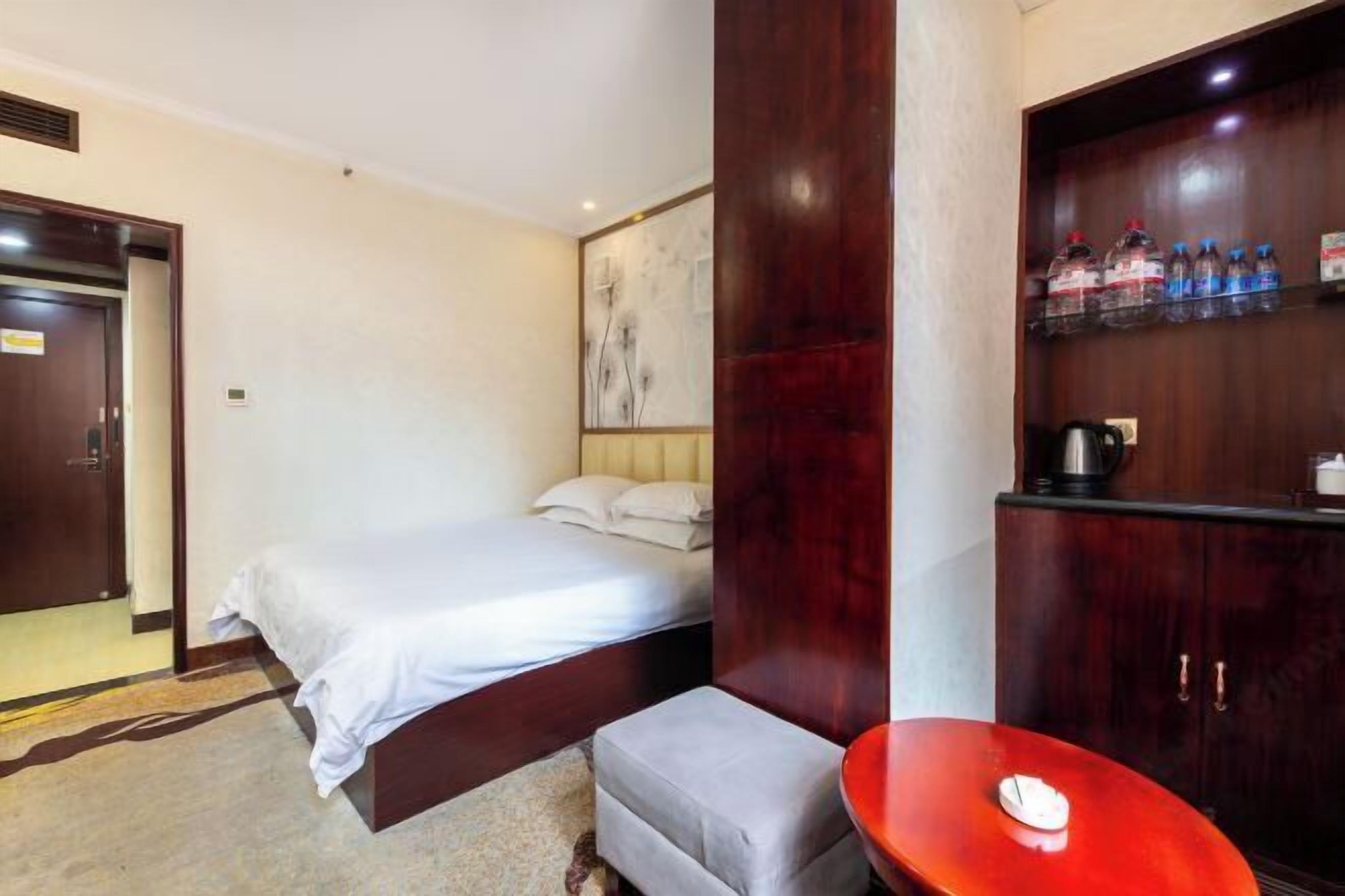 A hotel room with a bed, a small table, a cabinet with water bottles, and a painting on the wall.