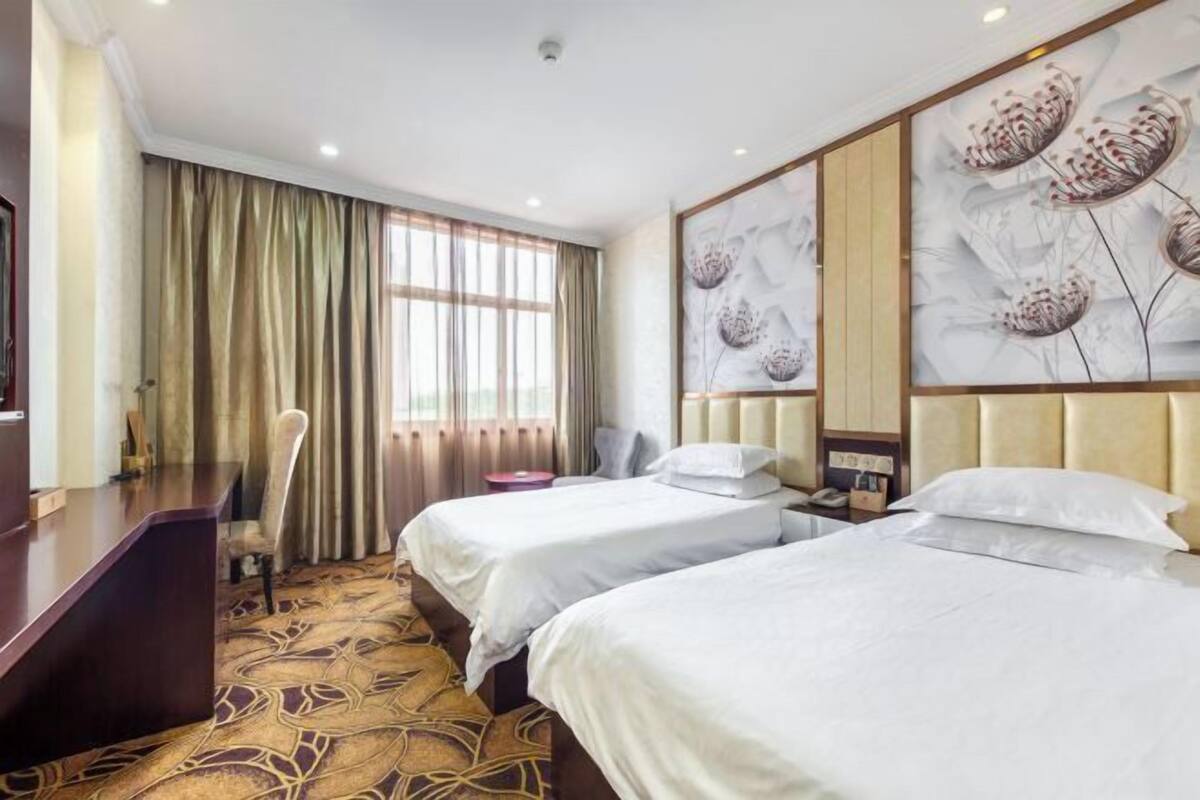 Habitación de hotel con dos camas, un escritorio, una silla y un amplio ventanal con cortinas.