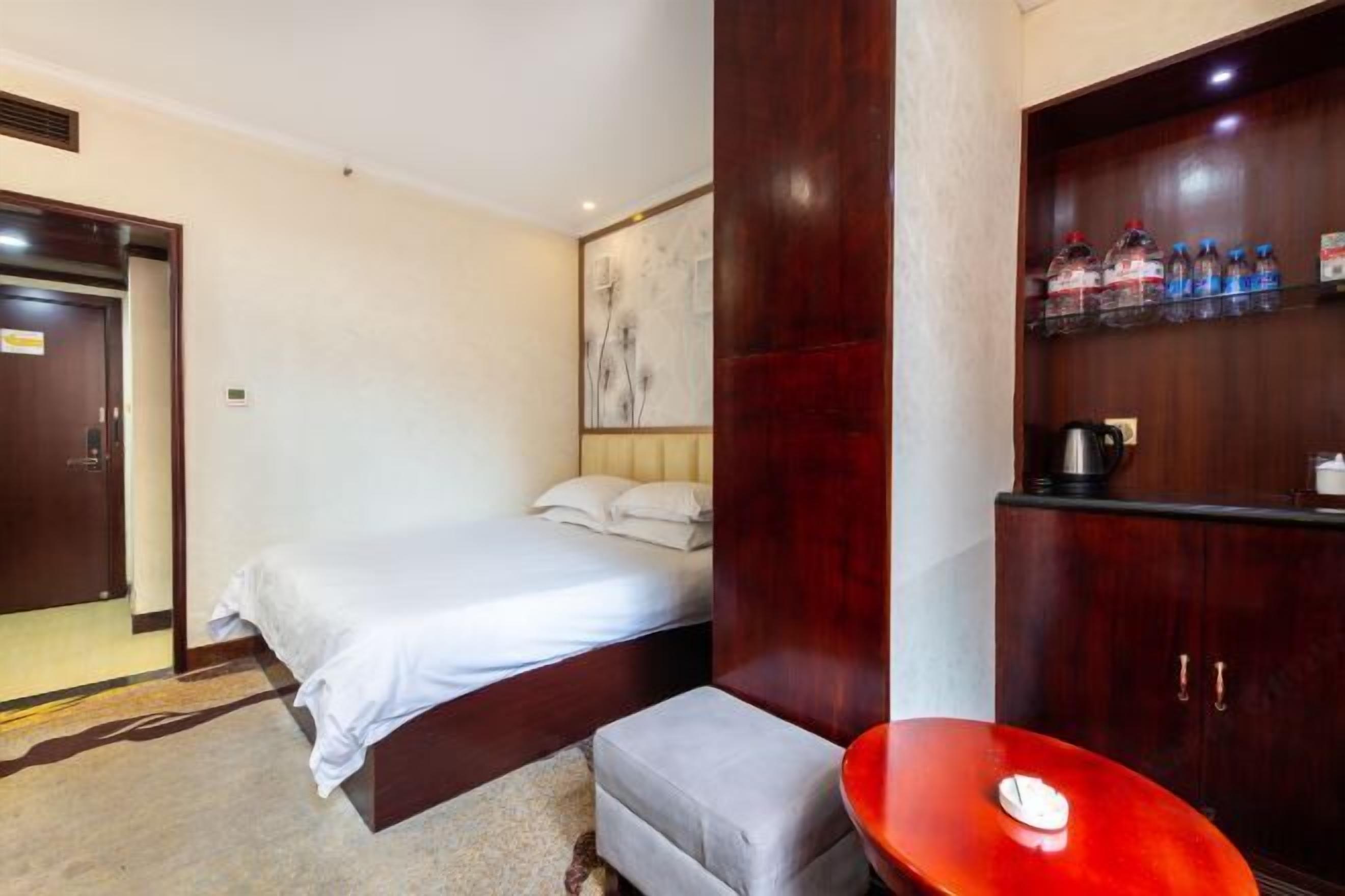 A hotel room with a bed, a small table, a cabinet with water bottles, and a painting on the wall.