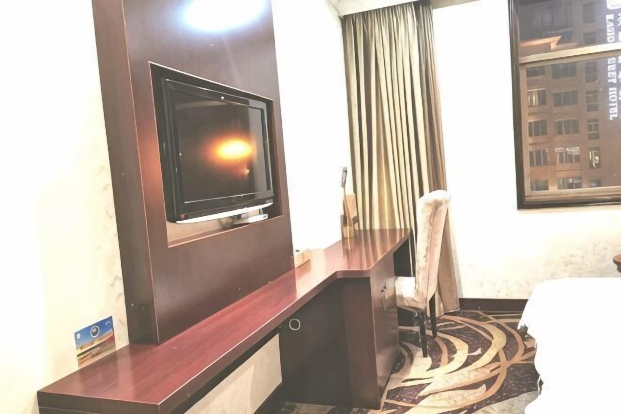 A hotel room with a television mounted on the wall, a desk with a chair, a bed, and a window with curtains.