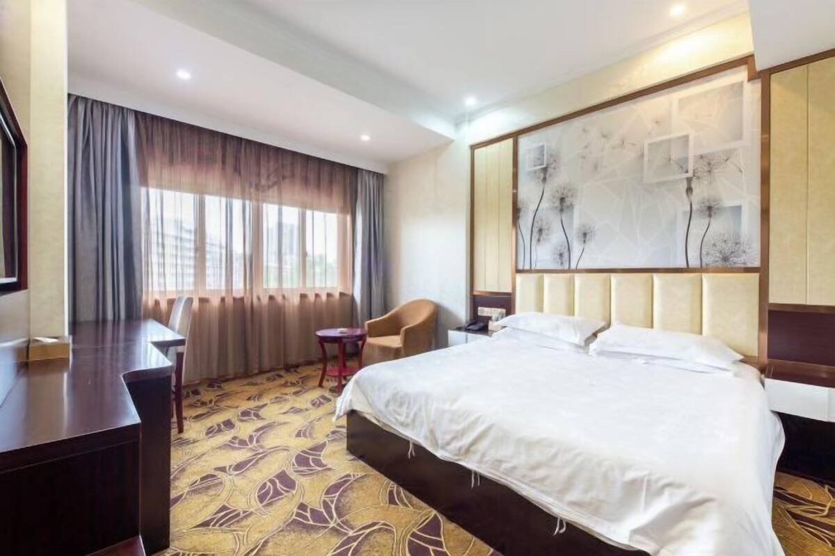 Habitación de hotel con una cama grande, un escritorio, una silla, una mesita y un ventanal con cortinas.
