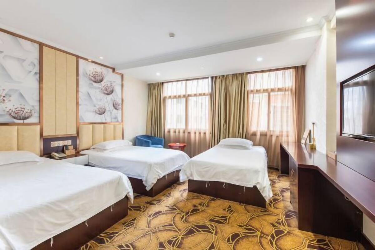 Habitación de hotel con tres camas, un televisor y un amplio ventanal con cortinas.