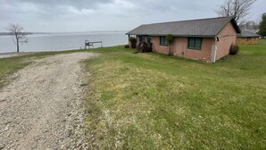 A single-story house with a gravel driveway leading to a wooden dock over a body of water.