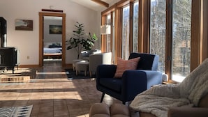 Una sala de estar acogedora con chimenea, asientos cómodos, un ventanal con vistas a árboles y un ventilador de techo.