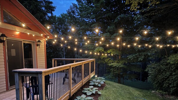 Una terraza con barandilla, rodeada por un jardín con plantas grandes y frondosas, bajo luces colgantes.