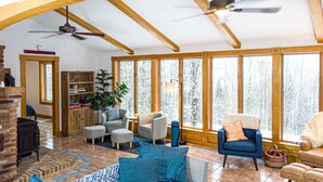 Amplia sala de estar con techo alto, vigas de madera, chimenea y grandes ventanales con vistas a un bosque nevado.