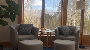 Una habitación con dos sillones y una mesita, ventanas amplias con vistas a árboles y montañas, y una lámpara sobre una mesita auxiliar.