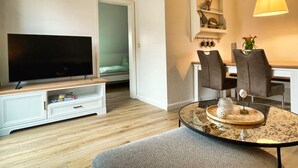 A living room with a flat-screen TV on a stand, a dining table with chairs, a small white cabinet, and a hanging lamp. - Schöne 3 Zi. Whg im Surflook in der Nähe der Familienlagune (Büsum)