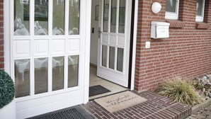 A house with a white front door, a brick exterior, and a small garden area with a welcome mat. - Schöne 3 Zi. Whg im Surflook in der Nähe der Familienlagune (Büsum)