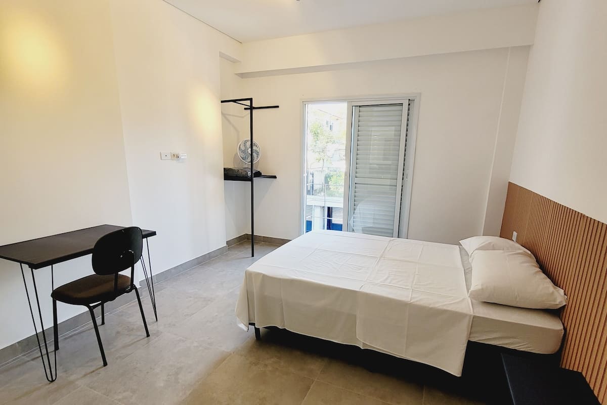 A modern bedroom with a bed, a desk, and a chair. There is a window with blinds, a ceiling fan, and a power outlet on the wall.