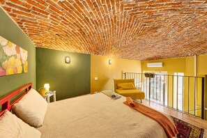 Une pièce avec un plafond orné d’un motif en briques, un lit avec une literie beige, un fauteuil jaune et un balcon doté d’une balustrade.