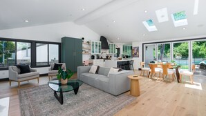 En rummelig opholdsstue med en grå sofa, et sofabord og et spisebord med stole. Der er et køkken i baggrunden med et stort vindue.