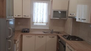 Cocina compacta con horno integrado, microondas y estufa a gas. Cuenta con alacenas de color claro y una ventana con cortinas.