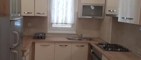 Cocina compacta con horno integrado, microondas y estufa a gas. Cuenta con alacenas de color claro y una ventana con cortinas.