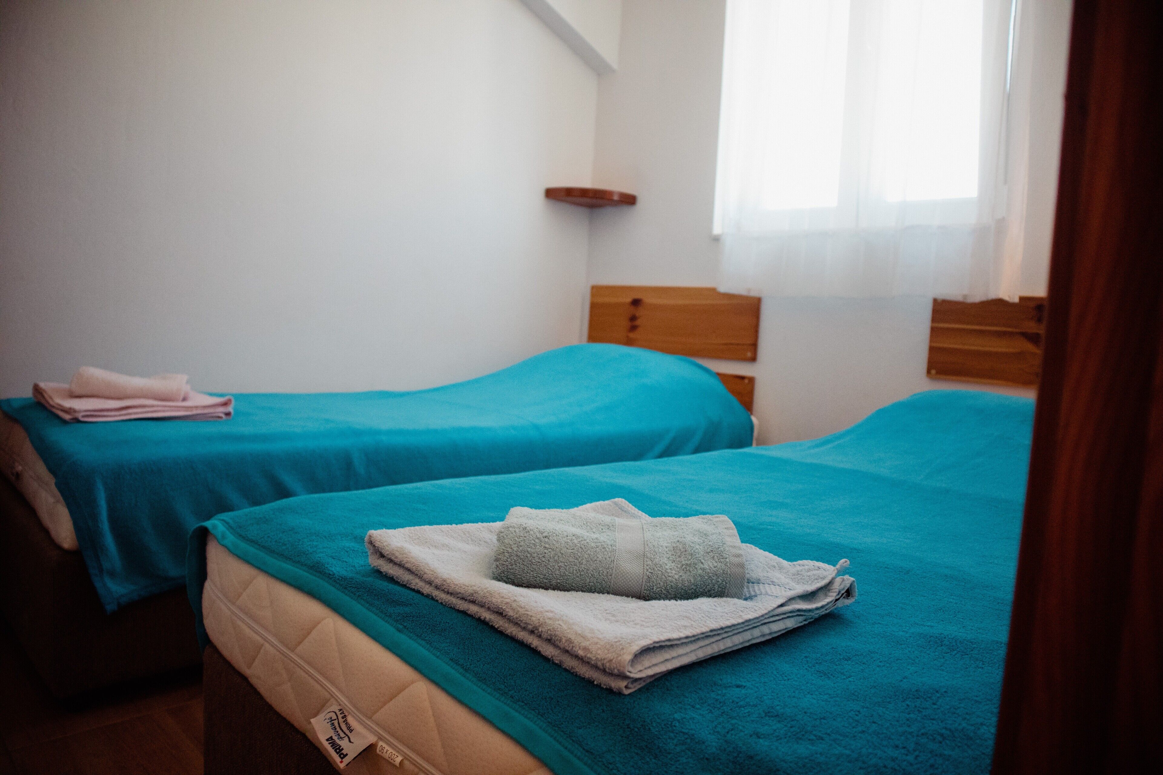 A room with two beds, each with a teal bedspread and a folded towel on the bed.
