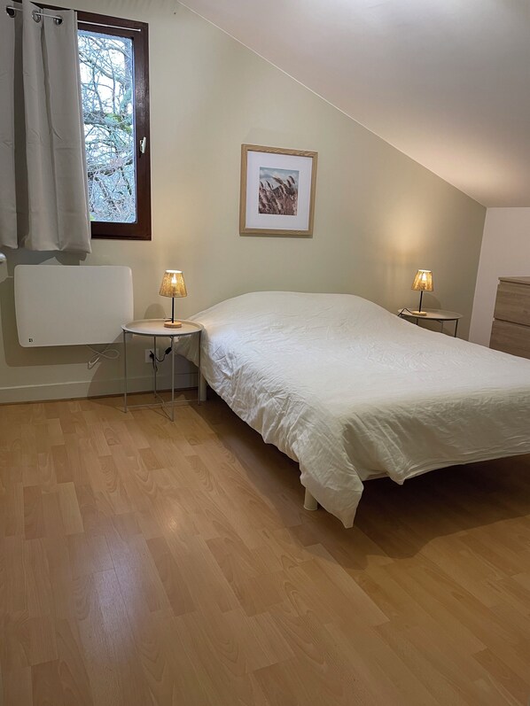A bedroom with a bed, two bedside tables with lamps, a window with curtains, a radiator, and a framed picture on the wall.