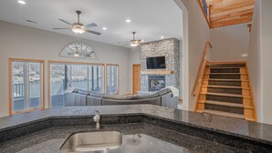 A spacious living area with a stone fireplace, a large window overlooking a body of water, and a modern kitchen with granite countertops. - Stunning Lake Life Vacation Home, 56 MM, spa, water toys, fire pit, dock/slip (Climax Springs)