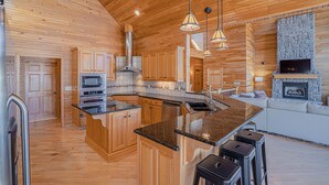 A spacious kitchen with wooden cabinetry, a central island, and a stone fireplace. - Stunning Lake Life Vacation Home, 56 MM, spa, water toys, fire pit, dock/slip (Climax Springs)