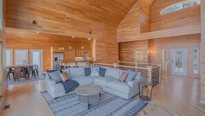 A spacious living area with a large L-shaped sectional sofa, a round coffee table, and a dining area with a kitchen in the background. - Stunning Lake Life Vacation Home, 56 MM, spa, water toys, fire pit, dock/slip (Climax Springs)