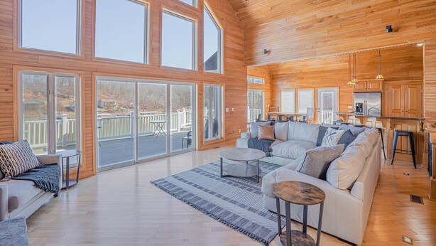 A spacious living room with a large sofa, a coffee table, and a rug. There are large windows and a deck area visible. - Stunning Lake Life Vacation Home, 56 MM, spa, water toys, fire pit, dock/slip (Climax Springs)