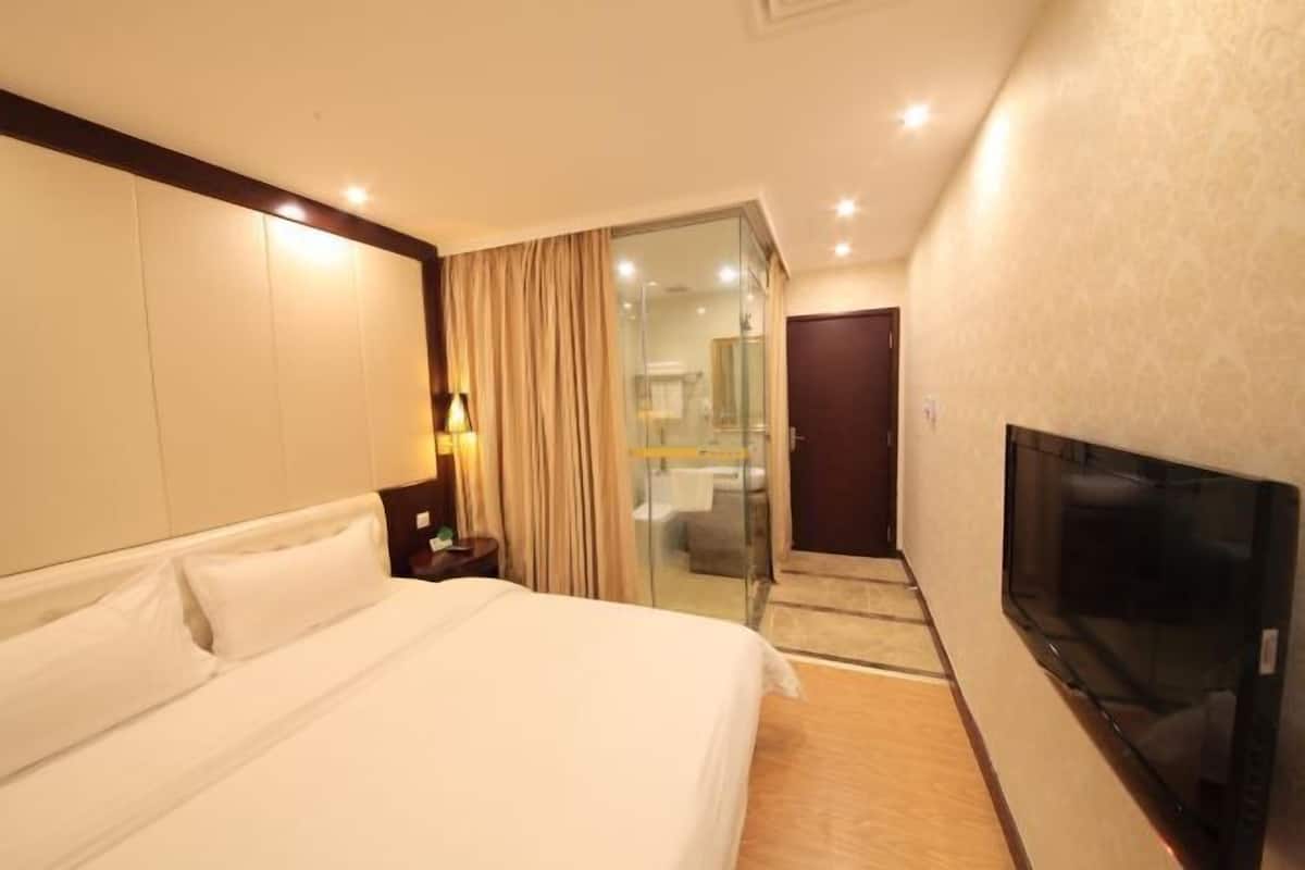A hotel room with a large bed, a TV, a glass door leading to a bathroom, and a small table with a lamp.