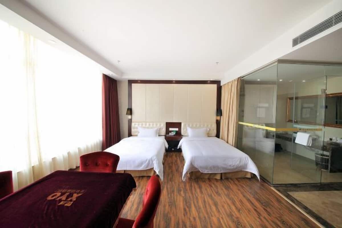 A hotel room with two beds, a red chair, a glass partition, and a view of a bathroom.