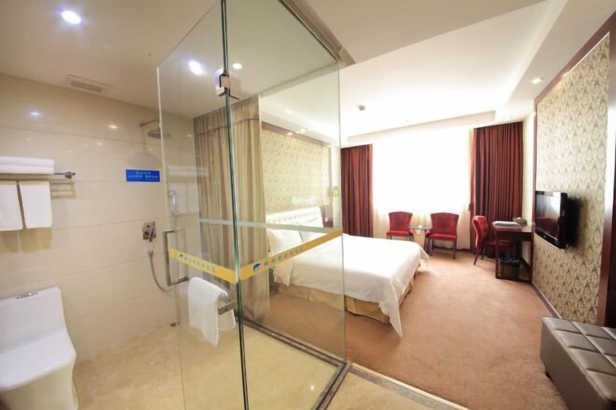 A hotel room with a glass partition, a bed, a desk, two chairs, a television, and a bathroom with a toilet and shower.