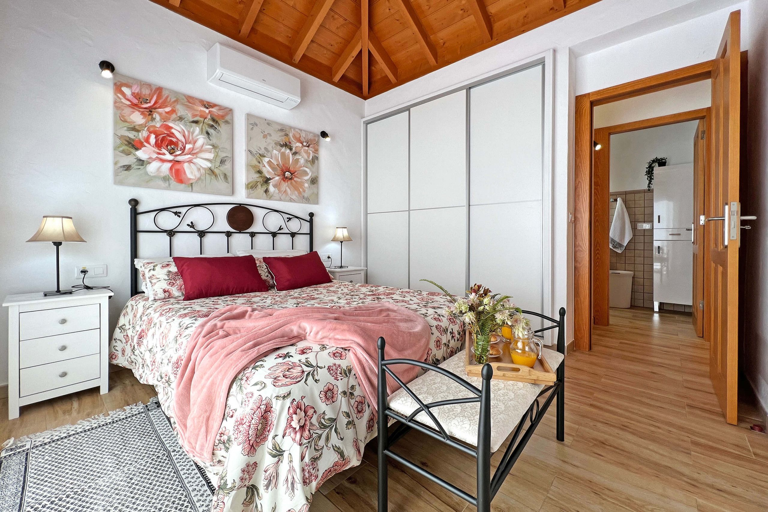 Un dormitorio con cama, mesitas de noche, un sillón, una mesa con un jarrón y techo de madera.