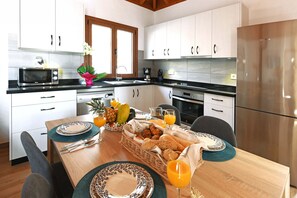 A modern kitchen with white cabinets, a stainless steel refrigerator, and a dining table set with plates, glasses, and a fruit basket. - Casas García, Tijarafe (Tijarafe)